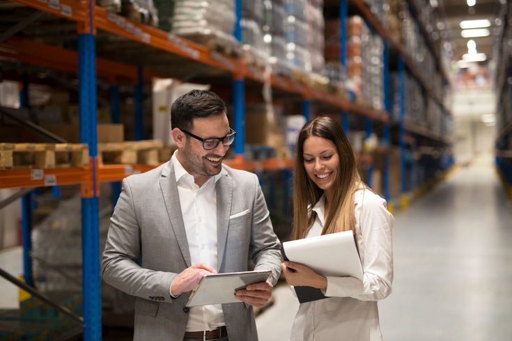 Dua profesional supply chain berdiskusi di gudang sambil memeriksa data di tablet dan dokumen sebagai bagian dari implementasi lean supply chain.
