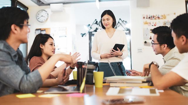 Tim profesional muda sedang berdiskusi di ruang kerja modern mengenai strategi manajemen sumber daya manusia. Seorang wanita berdiri memimpin diskusi sambil memegang tablet, sementara empat anggota tim lainnya duduk di sekitar meja dengan laptop, dokumen, dan catatan, terlibat aktif dalam perencanaan dan pengambilan keputusan terkait pengelolaan SDM