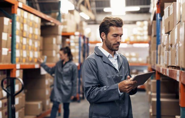 Seorang profesional supply chain memeriksa data inventaris menggunakan tablet di gudang sebagai bagian dari manajemen rantai pasok tersertifikasi.