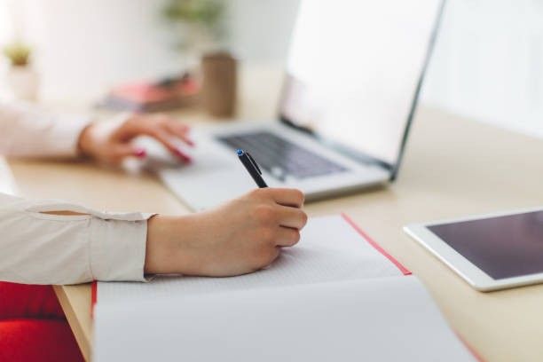 Seorang profesional sedang menulis di buku catatan sambil bekerja di depan laptop, menggambarkan proses belajar untuk mendapatkan Sertifikasi BNSP.